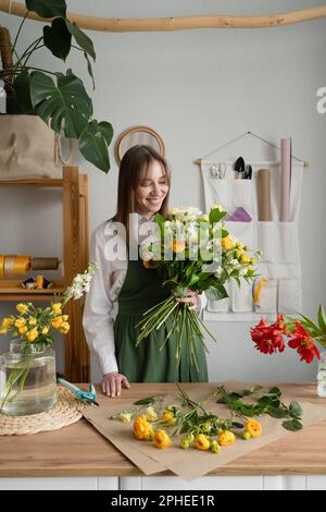 Weibliche Floristin in grüner Schürze steht am Tisch und macht während des Arbeitstages im Blumengeschäft einen Strauß frischer Blumen Stockfoto