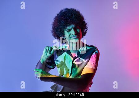 Junger lateinamerikanischer Mann mit Afro-Frisur und geschlossenen Augen, der ein Hemd trägt, während er im Studio tanzt und von violettem Neonlicht beleuchtet wird Stockfoto
