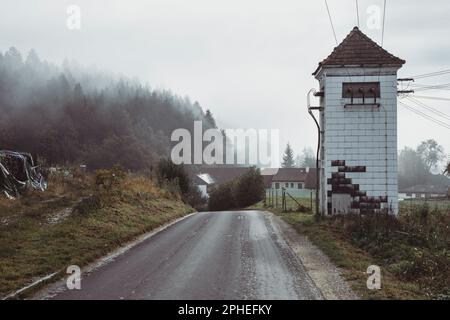 Leere gerade Asphaltstraße vorbei an halb zerstörten Ziegelseil Post und Wald auf Hügel unter bewölktem Himmel im Herbst Stockfoto