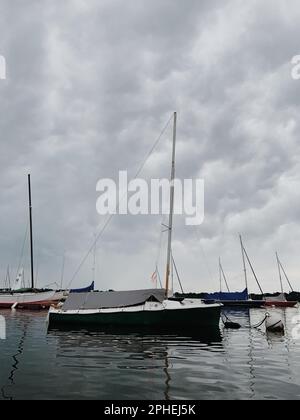 Eine Gruppe kleiner Segelboote wird auf einem See, der von einem wolkigen Himmel umgeben ist, schwimmend gesehen Stockfoto