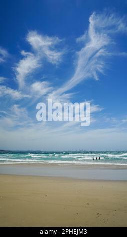 Surfstrand an einem windigen Tag. Blick vom Sandstrand über die Brandung bis zur Skyline der Stadt am Horizont, mit windgepeitschten Zirruswolken vor blauem Himmel. Stockfoto