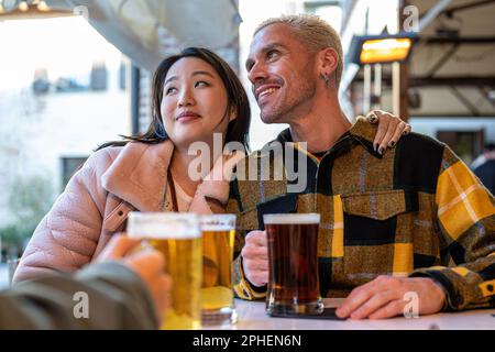 Zwei Freunde im Pub trinken Bier, ein junges, multiethnisches Paar mit Freunden im Pub, Konzept von Freundschaft, Liebe und kultureller Multiethnizität Stockfoto