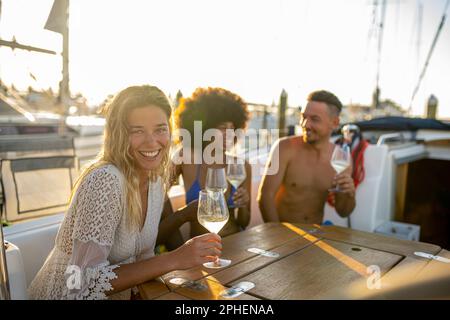 Partymomente auf einer Yacht, junge blonde Frau lächelt und schaut in die Kamera, Sonnenlicht durch die Segel der Boote im Hafen Stockfoto