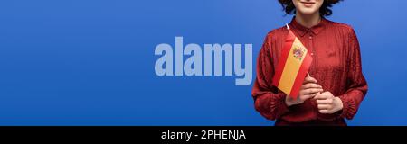 Zugeschnittener Blick auf einen glücklichen Sprachlehrer, der lächelt, während er die Flagge Spaniens isoliert auf blauem Banner hielt, Stockbild Stockfoto