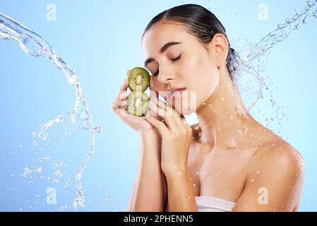 Frau, Wasserspritzer und Gesicht mit Kiwi in Relax für Hautpflege, Hygiene oder Ernährung vor blauem Studiohintergrund. Ruhige oder entspannte Haltung der Frau Stockfoto