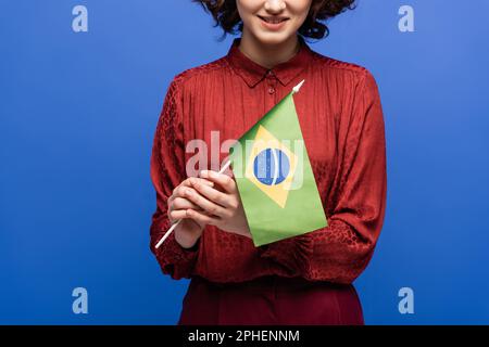 Zugeschnittener Blick auf einen glücklichen Sprachlehrer, der lächelt, während er die Flagge Brasiliens isoliert auf einem blauen Stockbild hält Stockfoto