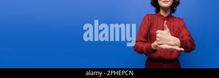 Zugeschnittene Ansicht einer fröhlichen Frau, die Hilfe-Geste auf Zeichensprache zeigt, isoliert auf blauem Banner, Stockbild Stockfoto