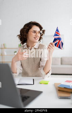 Fröhlicher Sprachlehrer, der auf die Flagge von Großbritannien schaut, während er zu Hause neben einem verschwommenen Laptop sitzt, Stock Image Stockfoto