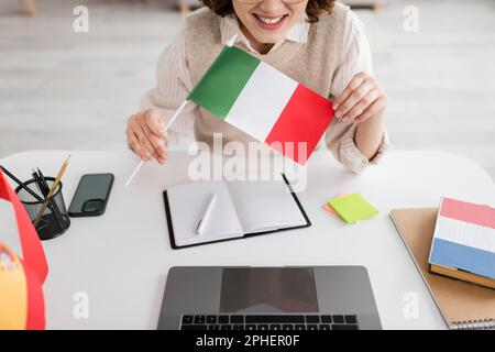 Zugeschnittene Ansicht eines lächelnden Sprachlehrers mit italienischer Flagge neben Geräten und Notebooks auf dem Tisch zu Hause, Stockbild Stockfoto