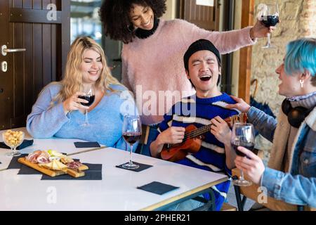 Internationale Gruppe von Freunden, die an einem Tisch in einem Pub im Freien sitzen, Spaß haben, singen und Rotwein trinken, inklusive und multiethnische Gruppe, asiatisch Stockfoto