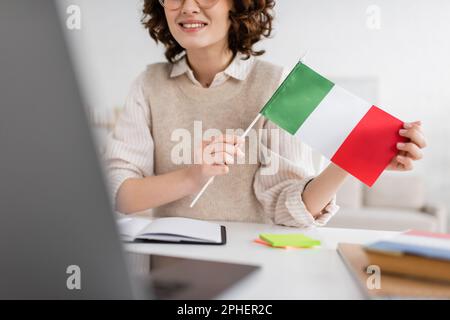 Teilansicht eines lächelnden Sprachlehrers mit der Flagge Italiens während des Online-Unterrichts auf verschwommenem Laptop zu Hause, Stockbild Stockfoto
