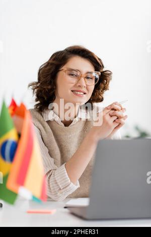 Fröhlicher Sprachlehrer in Brille neben Laptop und Flaggen auf verschwommenem Vordergrund, Stockbild Stockfoto