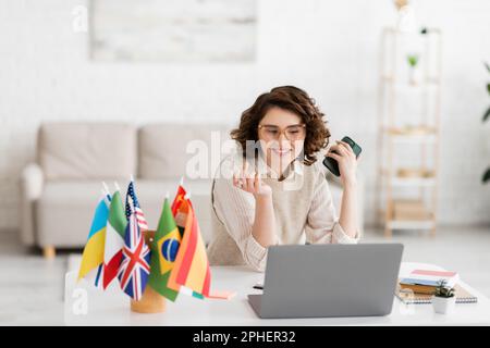 Fröhlicher Sprachlehrer in Brille hält Smartphone in der Nähe des Laptops und verschiedene Flaggen auf verschwommenem Vordergrund, Stockbild Stockfoto