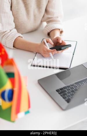 Zugeschnittene Ansicht eines Sprachlehrers mit Smartphone in der Nähe eines Laptops und Flaggen auf verschwommenem Vordergrund, Stockbild Stockfoto