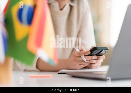 Zugeschnittene Ansicht eines Sprachlehrers, der ein Mobiltelefon in der Nähe eines Laptops verwendet, und Flaggen auf verschwommenem Vordergrund, Stockbild Stockfoto