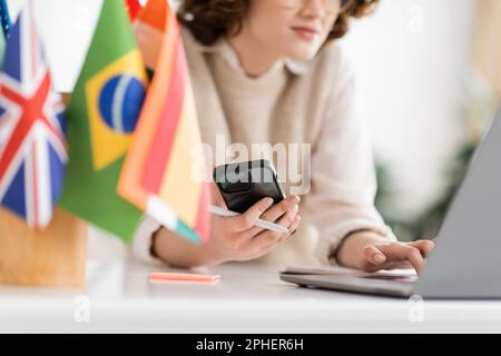 Zugeschnittene Ansicht eines Sprachlehrers, der ein Mobiltelefon in der Nähe eines Laptops verwendet, und verschiedene Flaggen auf verschwommenem Vordergrund, Stockbild Stockfoto