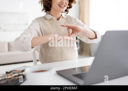 Zugeschnittene Ansicht eines fröhlichen Lehrers, der während des Online-Unterrichts eine Geste der Gebärdensprache auf dem Laptop zeigt, Stockbild Stockfoto