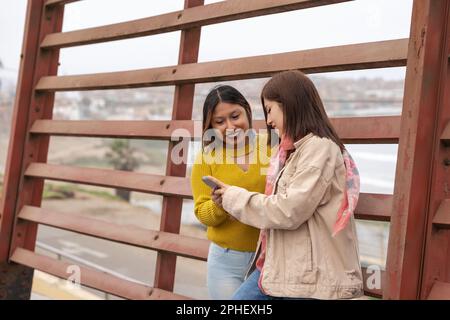 Zwei junge lateinische Freundinnen stehen auf der Straße und benutzen ein Handy Stockfoto