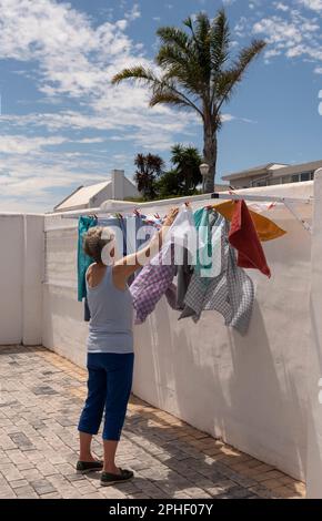 Langebaan, Südafrika. 2023. Ältere Frau, die an einem windigen Tag in ihrem Garten die Wäsche aufhängt. Stockfoto