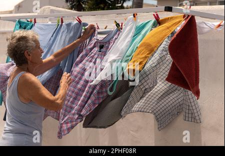 Langebaan, Südafrika. 2023. Ältere Frau, die an einem windigen Tag in ihrem Garten die Wäsche aufhängt. Stockfoto