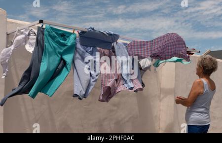 Langebaan, Südafrika. 2023. Ältere Frau, die an einem windigen Tag in ihrem Garten die Wäsche aufhängt. Stockfoto