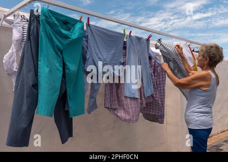 Langebaan, Südafrika. 2023. Ältere Frau, die an einem windigen Tag in ihrem Garten die Wäsche aufhängt. Stockfoto