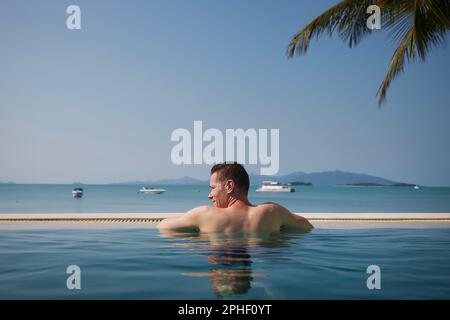 Mann mit Blick nach hinten, während man sich im Swimmingpool gegen das Meer mit Inseln entspannt. Urlaub am Strand in tropischem Reiseziel. Stockfoto