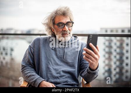Ein hübscher älterer Mann mit grauen Haaren und Brille, der auf der Terrasse sitzt, Zigaretten und Smartphones benutzt, kommuniziert, Spaß hat und Spaß hat Stockfoto