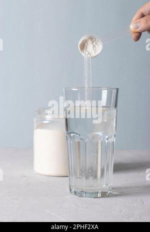 Frau fügt einem Glas Wasser mit einem Messlöffel Kollagenpulver hinzu. Gesundes und Anti-Age-Konzept auf blauem Hintergrund Stockfoto