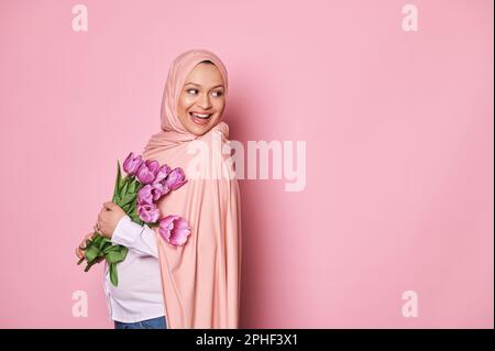 Eine wunderschöne muslimische Frau in pinkfarbenem Hijab, die einen Strauß Tulpen hält und lächelt, während sie auf den Kopierbereich auf pinkfarbenem Hintergrund blickt Stockfoto