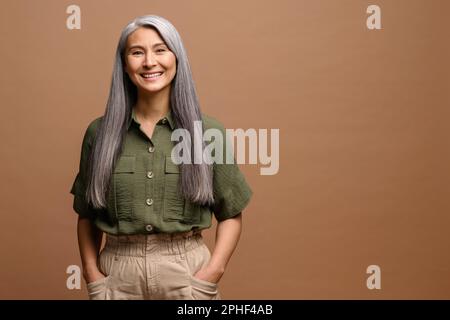Fröhliches Lächeln stilvoll selbstbewusst 50 Jahre alte asiatische wunderschöne Frau, die auf den braunen Hintergrund in die Kamera schaut. Grauhaarige Frau mittleren Alters isoliert, Kopierraum Stockfoto