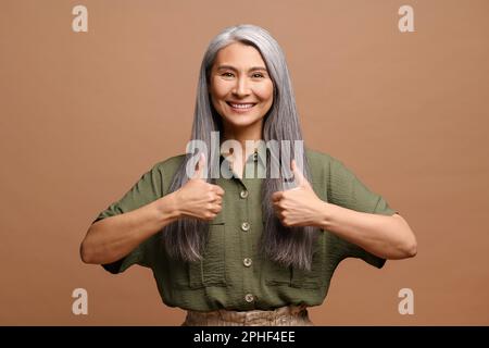 Attraktive 50s-jährige Frau mittleren Alters mit langen grauen Haaren, die Daumen nach oben zeigt und mit einem zahnigen Lächeln in die Kamera schaut, eine Dame, die Ihnen zustimmt, isoliert auf braun, gute Kritik Stockfoto