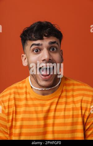 Junger, attraktiver, stilvoller Mann mit offenem Mund und durchstechender Halskette, der auf einem isolierten orangefarbenen Hintergrund steht Stockfoto