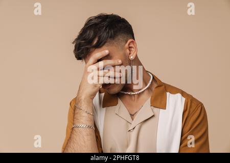Porträt eines jungen, stilvollen Mannes mit Halskette, der sein Gesicht mit der Hand bedeckt, während er über einem isolierten braunen Hintergrund steht Stockfoto