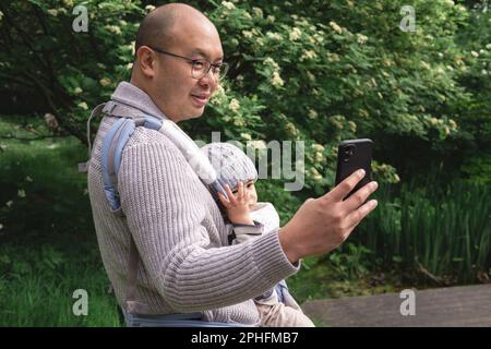 Ein lächelnder neuer Vater, der sein männliches Kind mit einer Babytragetasche trägt und im Frühling mit seinem Handy ein Selfie-Foto in der Natur im Park macht. Briti Stockfoto