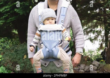 Ein nicht wiedererkennbarer Elternteil, der sein männliches Kind mit einem Babyträger trägt und im Frühling in der Natur im Park unterwegs ist. Der Sohn hat eine gelbe Blume Stockfoto