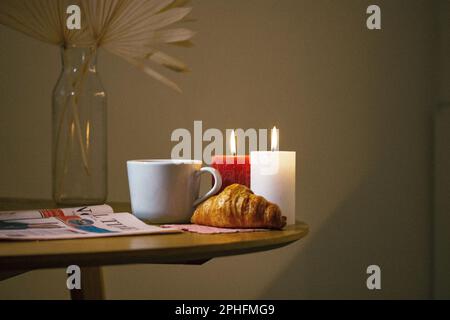 Eine weiße Keramiktasse Tee oder Kaffee steht auf einem Holztisch neben einem frisch gebackenen Croissant und zwei brennenden Kerzen. Ein gemütliches beigefarbenes Stillleben mit einer vier Stockfoto