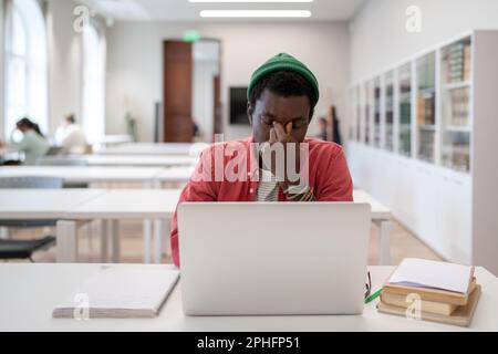 Ein müder Schwarzer massiert die Nase, sitzt mit geschlossenen Augen vor dem Laptop in der Bibliothek Stockfoto