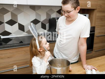 Bruder mit Brille und seine kleine Schwester in der Küche, die beim Kochen lächelt und sich ansieht. Kindermischteig in Hasenohren. Helft mir, Kumpel Stockfoto