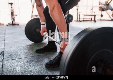 Die Klingel wird angehoben. Der junge sportliche, starke Mann in schwarzer Kleidung trainiert im Fitnessstudio Stockfoto
