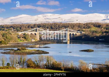 Malerischer Blick auf die Menai-Hängebrücke, die die Menai-Straße überquert, mit Schnee auf den Bergen im Winter. Menai Bridge, Isle of Anglesey, Wales, Vereinigtes Königreich Stockfoto
