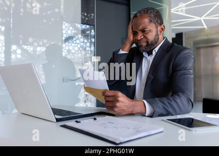 Ein nervöser und aufgebrachter Geschäftsmann im Büro erhielt eine Nachricht mit einer Nachricht über einen Briefumschlag mit schlechten Nachrichten, Bankrott, ein afroamerikanischer Mann traurig und nachdenklich, der ein Dokument las. Stockfoto