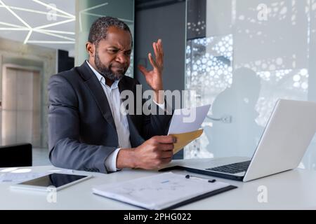 Ein nervöser und aufgebrachter Geschäftsmann im Büro erhielt eine Nachricht mit einer Nachricht über einen Briefumschlag mit schlechten Nachrichten, Bankrott, ein afroamerikanischer Mann traurig und nachdenklich, der ein Dokument las. Stockfoto