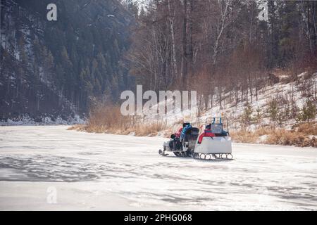 Winterspaziergang mit der Familie auf einem Schneemobil auf einem gefrorenen Fluss. Ein Schneemobil mit Anhänger rollt Kinder auf dem Eis des eisigen Sibirien Mana River in Stockfoto