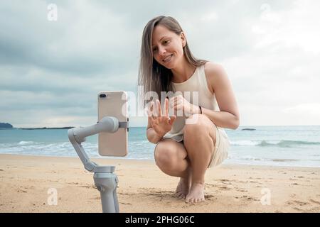 Eine Bloggerin erschießt sich in einem Video-Blog am Meer mit einem Handy. Eine junge Frau winkt und lächelt in der Begrüßung, während sie sich auf einem SM filmt Stockfoto
