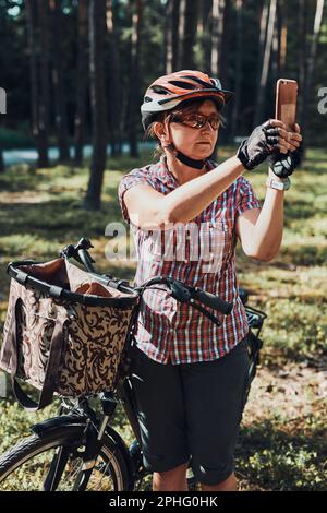 Aktive Frau verbringt kostenlose Sommerferien in einem Wald auf einer Fahrradtour und macht Fotos mit dem Smartphone. Frau mit Fahrradhelm und Handschuhen Stockfoto