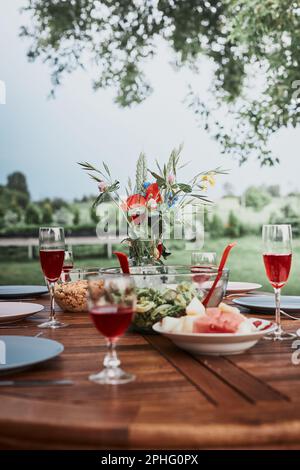 Abendessen in einem Apfelgarten auf Holztisch mit Salaten und Wein mit Blumen dekoriert. Nahaufnahme des Tisches mit Essen für Familienessen vorbereitet Stockfoto