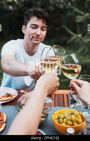 Freunde, die während des Picknick-Abendessens im Freien in einem heimischen Garten anstoßen. Nahaufnahme von Leuten, die Weingläser mit Weißwein über den Tisch mit piz halten Stockfoto