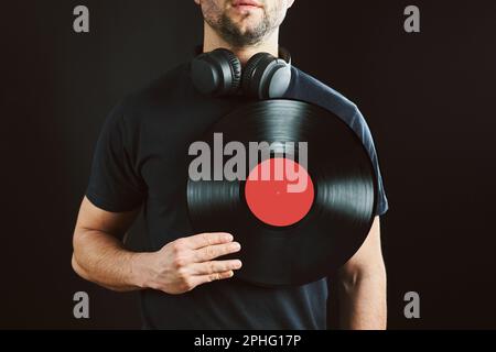 Ein Mann, der eine Schallplatte über dem Herzen hält. Leidenschaft für Musik. Klassischer Musikstil. Männlich mit Kopfhörern mit alter Vinylscheibe auf schwarzem Hintergrund. Ret Stockfoto