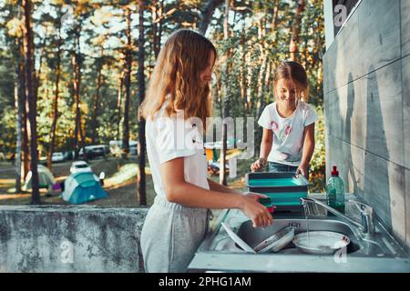 Teenager-Mädchen waschen das Geschirr Töpfe und Teller mit Hilfe ihrer jüngeren Schwester in der Outdoor-Küche während des Urlaubs auf Camping. Lagerleben. Schwester Stockfoto